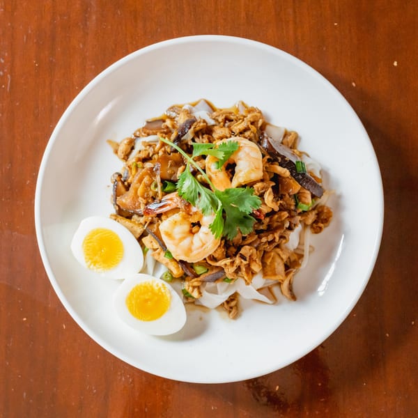 A white plate of stir-fried noodles with shrimp, mushrooms, and two halved boiled eggs.
