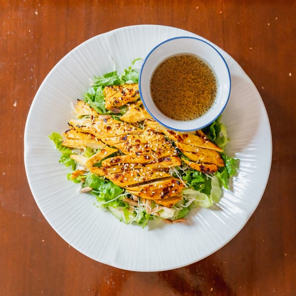 Grilled chicken salad with sesame seeds, greens, and a side of dressing on a white plate.