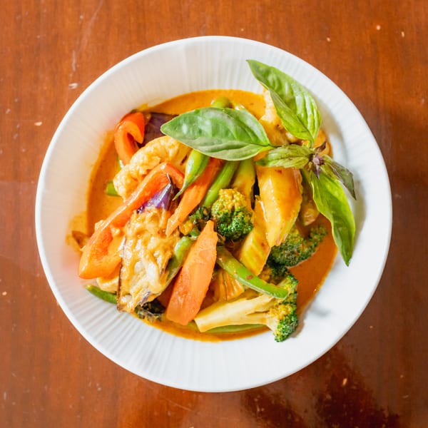 Top-down view of a white bowl filled with red curry, vegetables, and basil leaves.