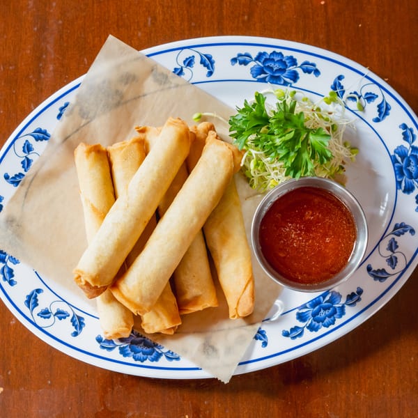 Crispy golden spring rolls with sweet chili sauce, parsley, and bean sprouts on a blue and white plate.